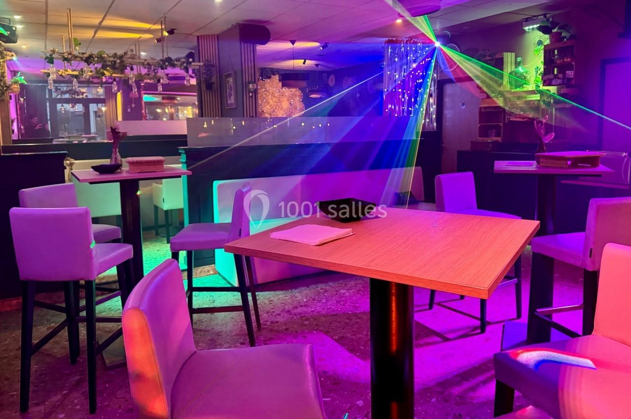Salle de bar avec tables hautes, chaises, éclairage coloré et effets lumineux dans une ambiance festive.