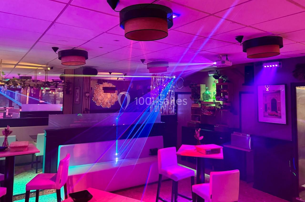 Salle de bar ou restaurant avec éclairage coloré, tables hautes et faisceaux lumineux créant une ambiance festive.