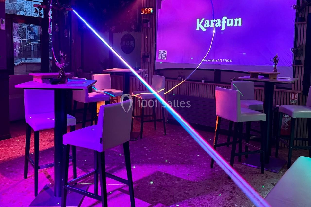 Salle de karaoké avec éclairage coloré, tables hautes, chaises blanches et écran affichant ’Karafun’.