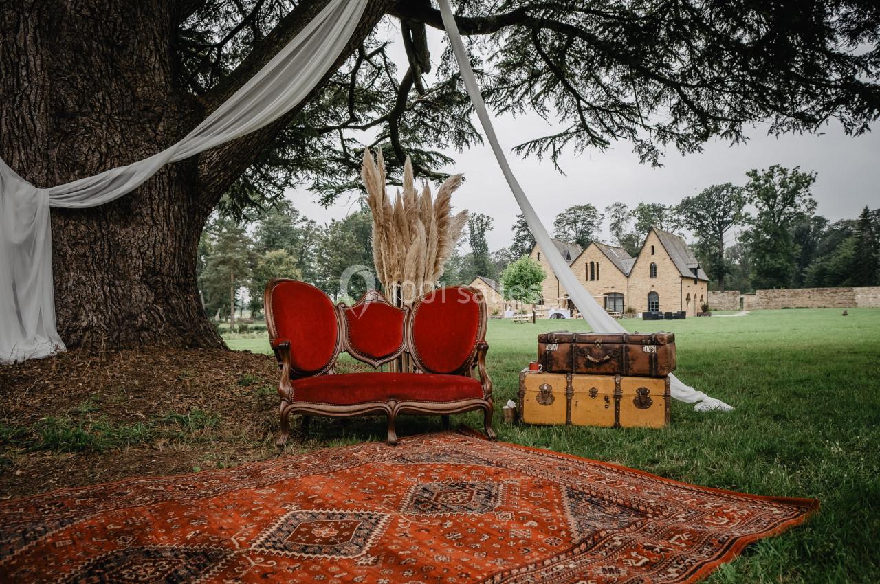 Canapé rouge, tapis oriental, valises anciennes et rideaux blancs sous un grand arbre, avec une maison en pierre au fond.