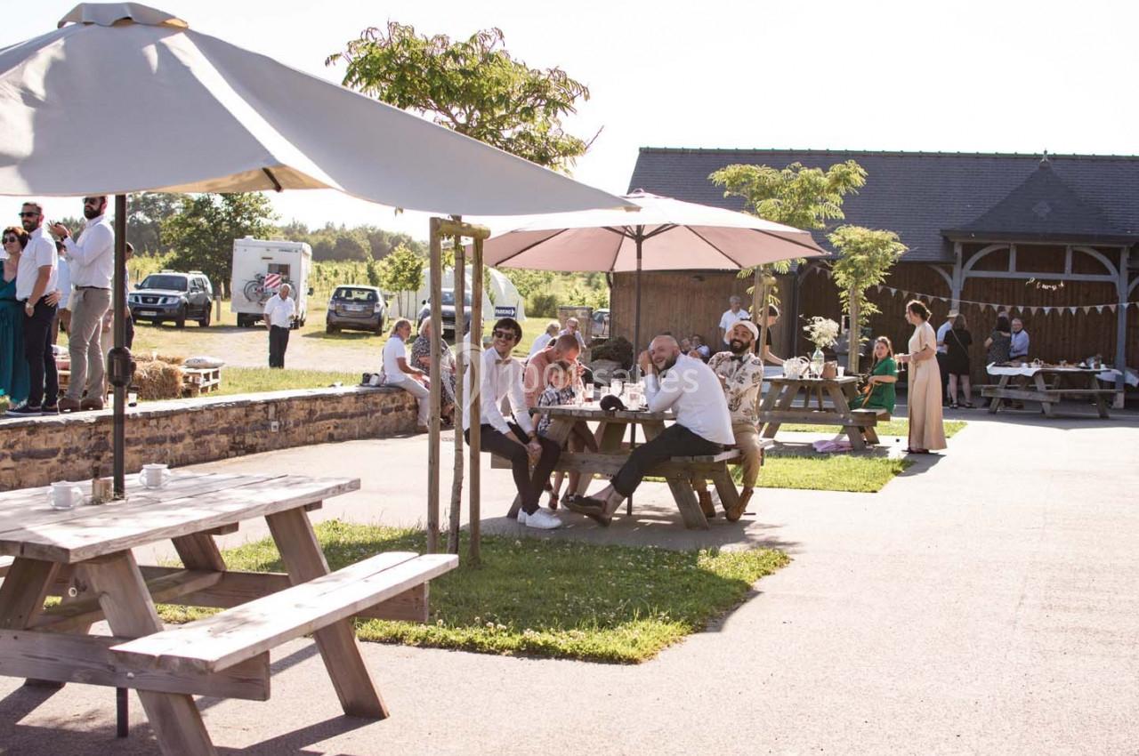 Personnes assises à des tables en bois sous des parasols dans une cour ensoleillée près d'un bâtiment en pierre.