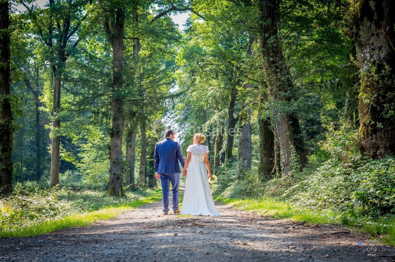 Un couple marche main dans la main sur un chemin forestier bordé d'arbres sous une lumière naturelle.