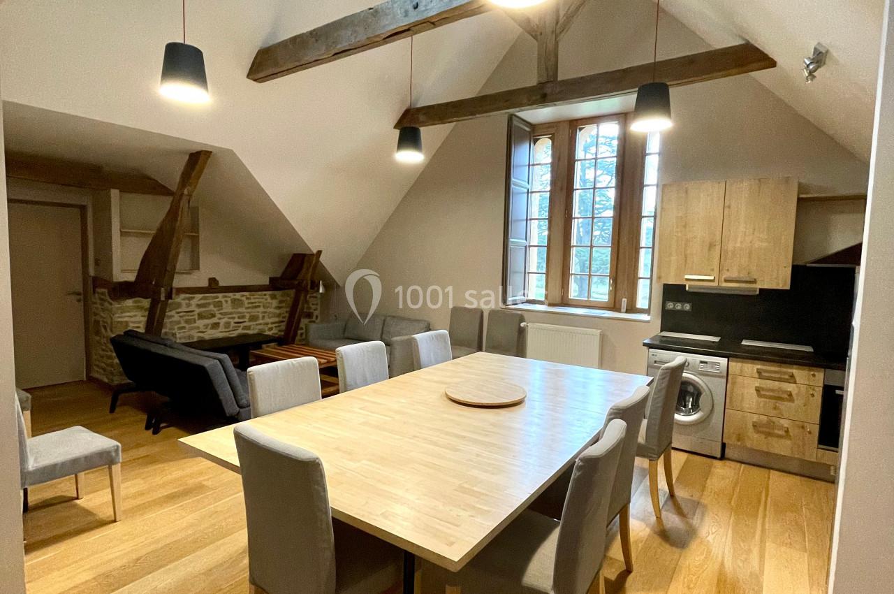 Salle à manger lumineuse avec table en bois, chaises grises, cuisine équipée et poutres apparentes sous un plafond mansardé.