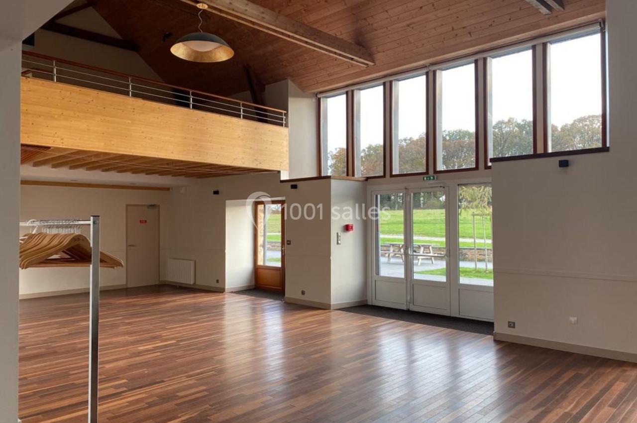 Salle lumineuse avec parquet en bois, grande baie vitrée, mezzanine et portants à vêtements sur la gauche.