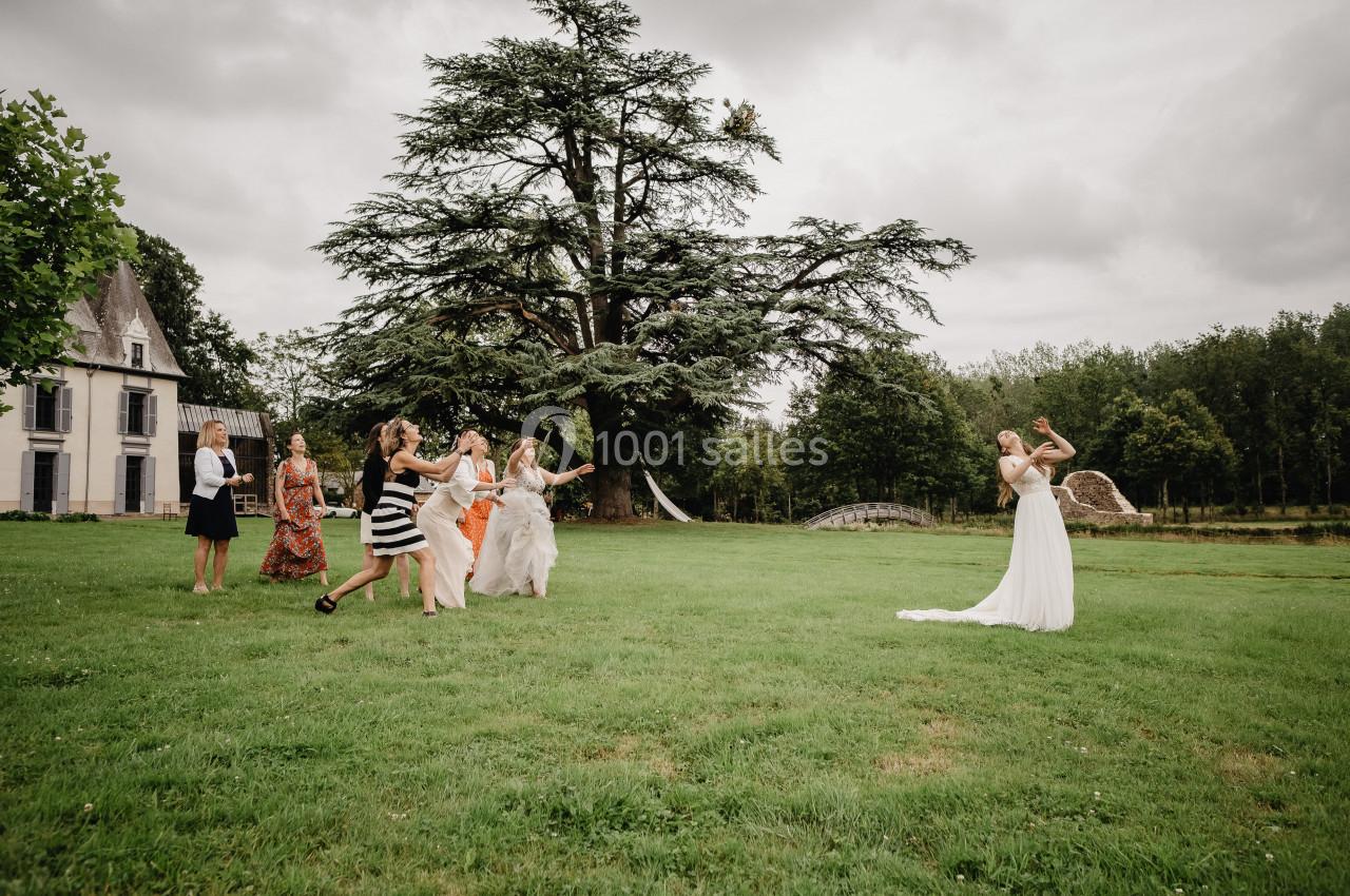 Une mariée lance son bouquet à un groupe de femmes sur une pelouse devant un grand arbre et un bâtiment.