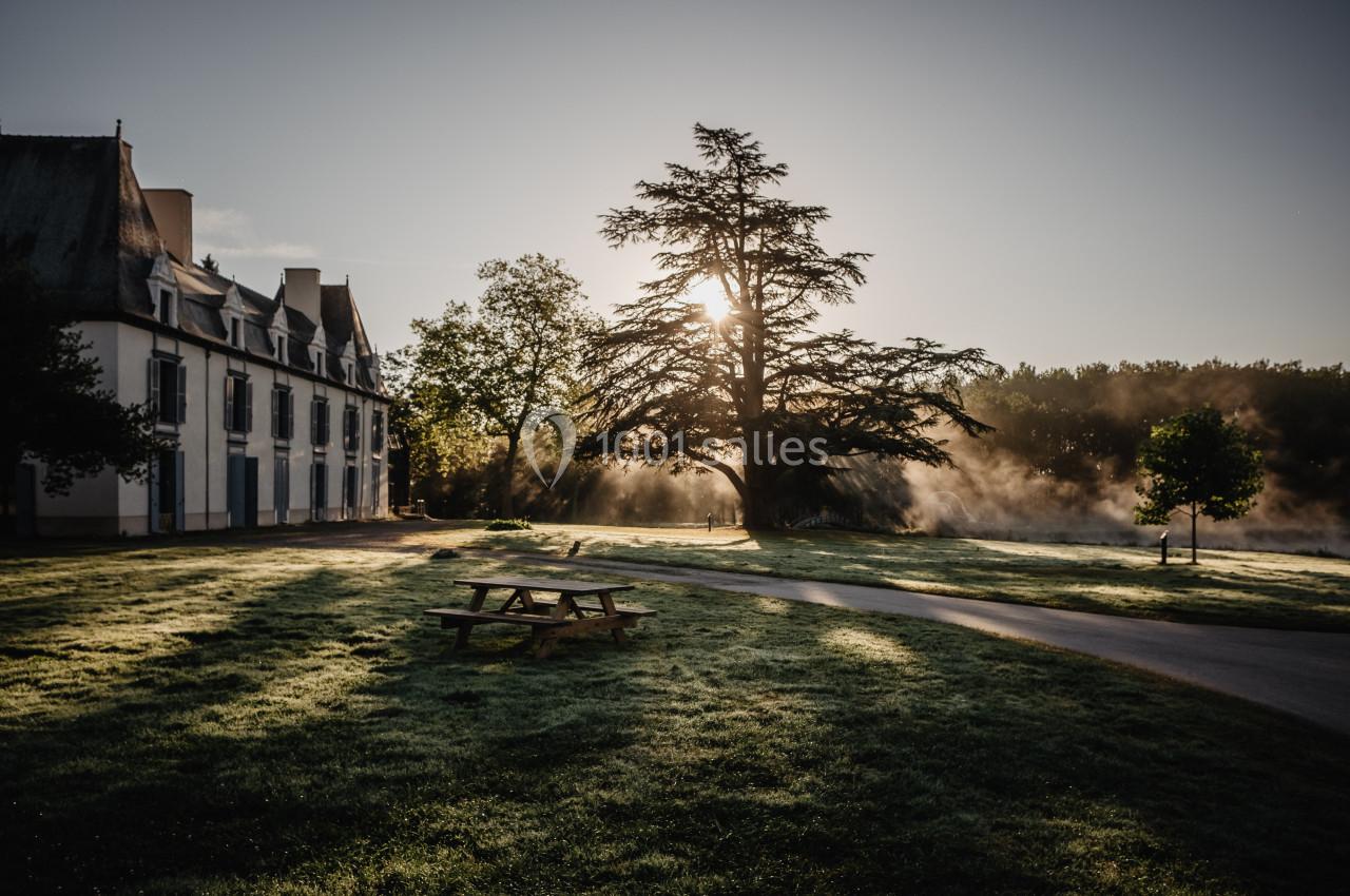 Pelouse au lever du soleil avec un grand arbre, une maison en pierre et des bancs de pique-nique dans un cadre paisible.
