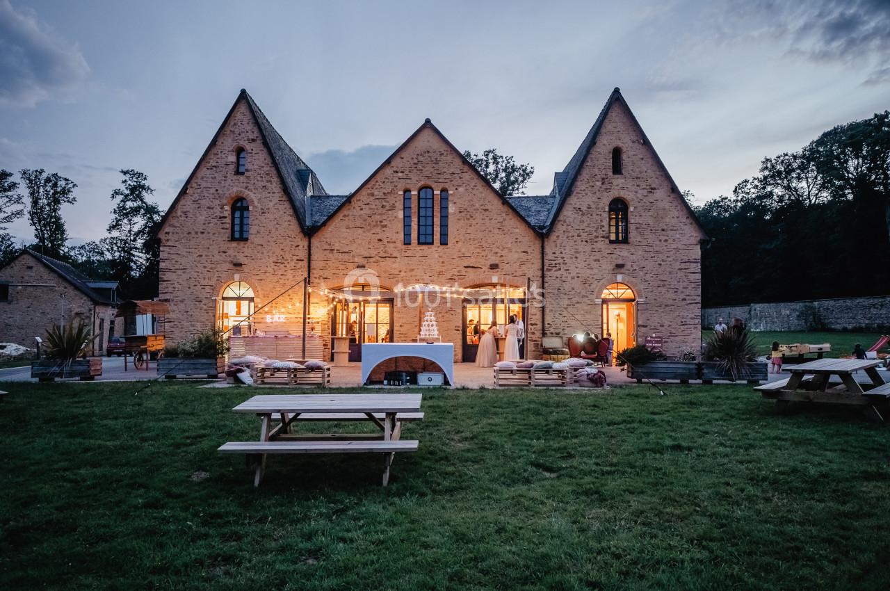 Bâtiment en pierre avec grandes fenêtres éclairées au crépuscule, entouré de pelouses et de tables de pique-nique.