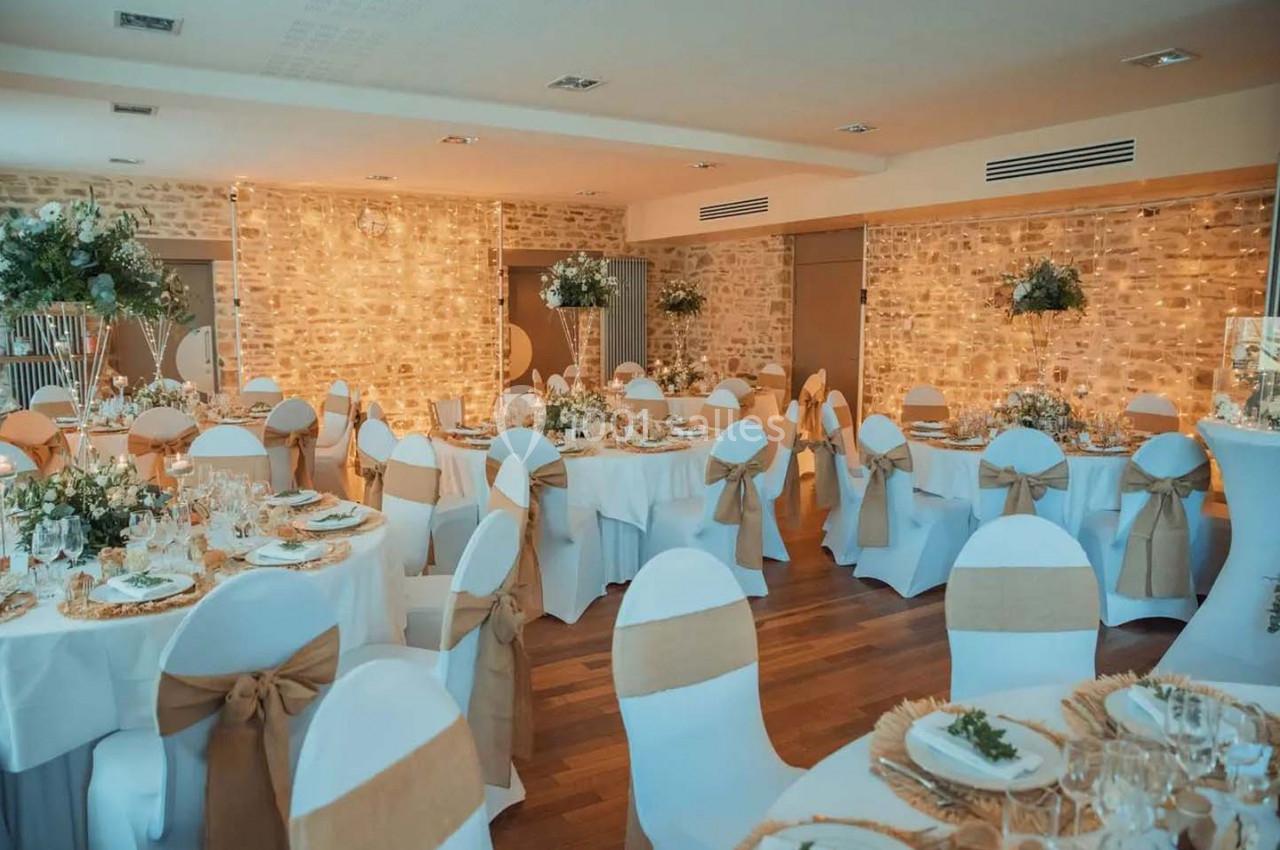 Salle de réception décorée avec des tables rondes, nappes blanches, chaises ornées de nœuds beige et guirlandes lumineuses.