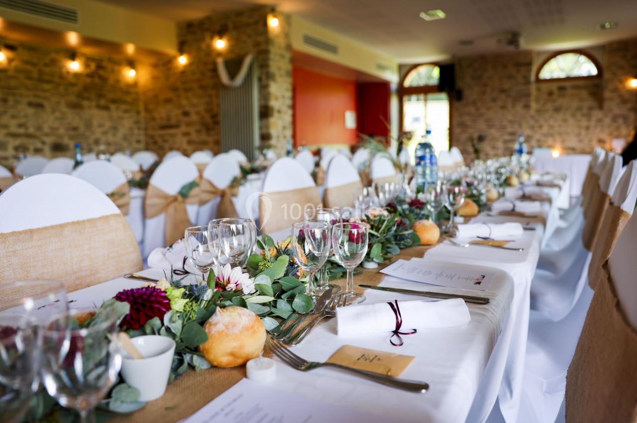 Salle de réception décorée avec des tables alignées, nappes blanches, centres de table fleuris et chaises ornées de toile de…