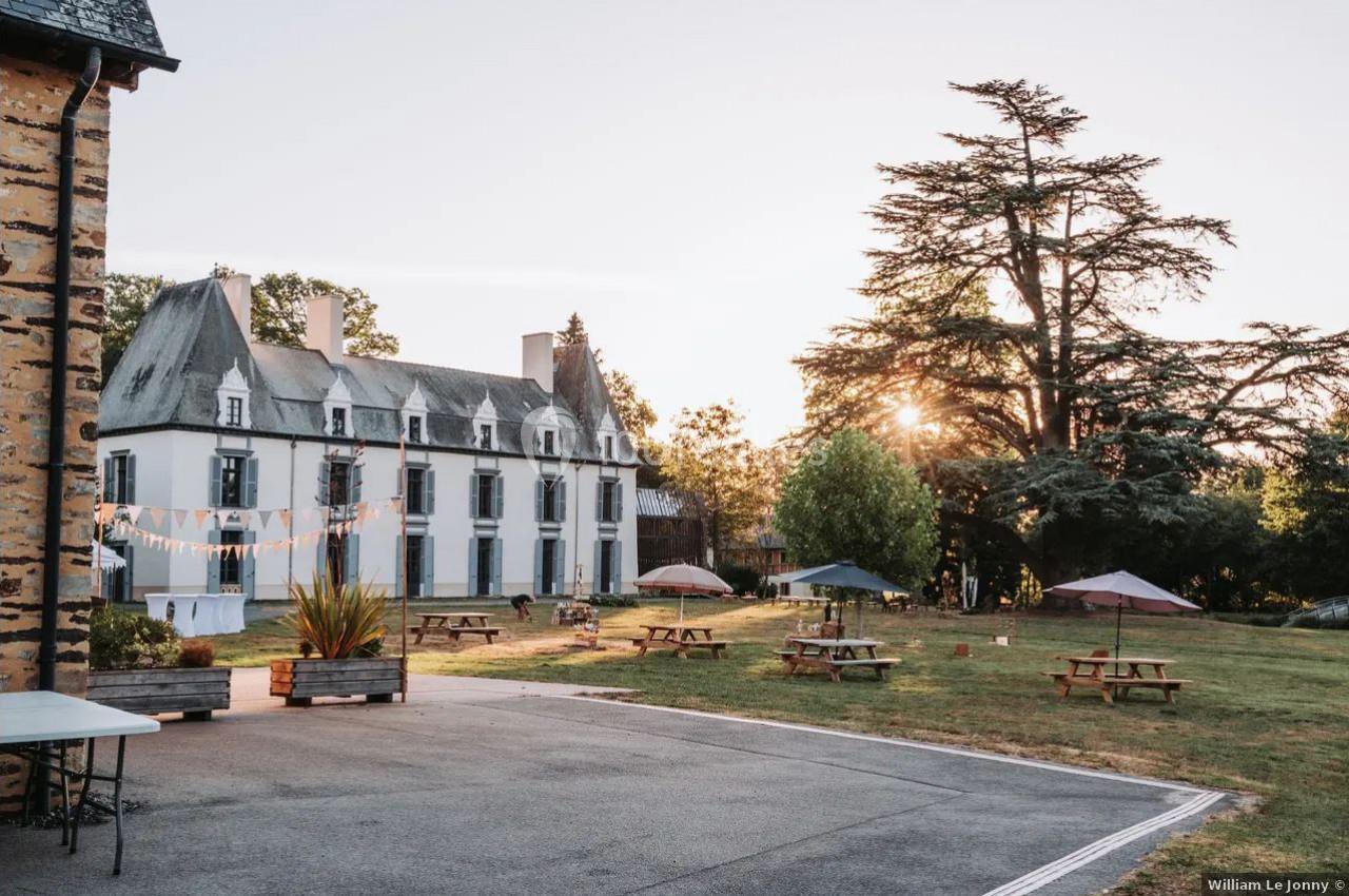 Cour d’un château avec pelouse, tables de pique-nique, parasols et arbres, éclairée par un coucher de soleil.