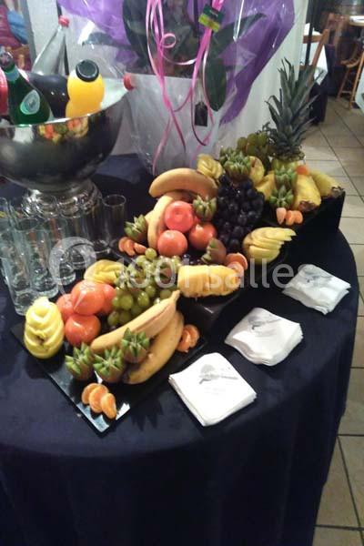 Plateau de fruits variés avec ananas, bananes, raisins et mandarines, disposé sur une table avec serviettes et boissons.