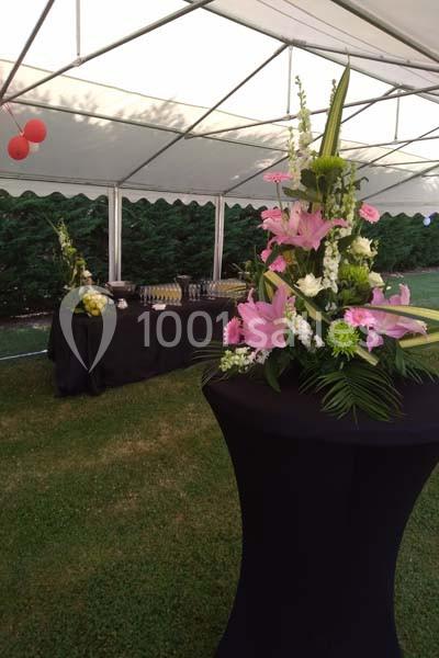 Arrangement floral avec des lys roses sur une table haute, sous une tente blanche, avec un buffet en arrière-plan.