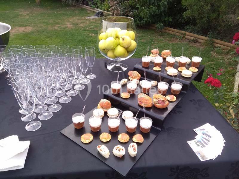 Table dressée avec des verrines, amuse-bouches, verres vides et une coupe remplie de pommes vertes, en extérieur.