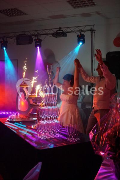 Un couple de mariés célèbre devant une fontaine de verres illuminée, entouré de lumières colorées et de feux d'artifice.