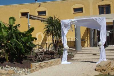 Cour extérieure avec végétation tropicale, terrasse ensoleillée, murs jaunes et mobilier en bois près d'une pergola.