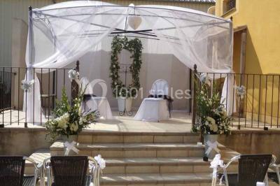 Cour extérieure avec végétation tropicale, terrasse ensoleillée, murs jaunes et mobilier en bois près d'une pergola.