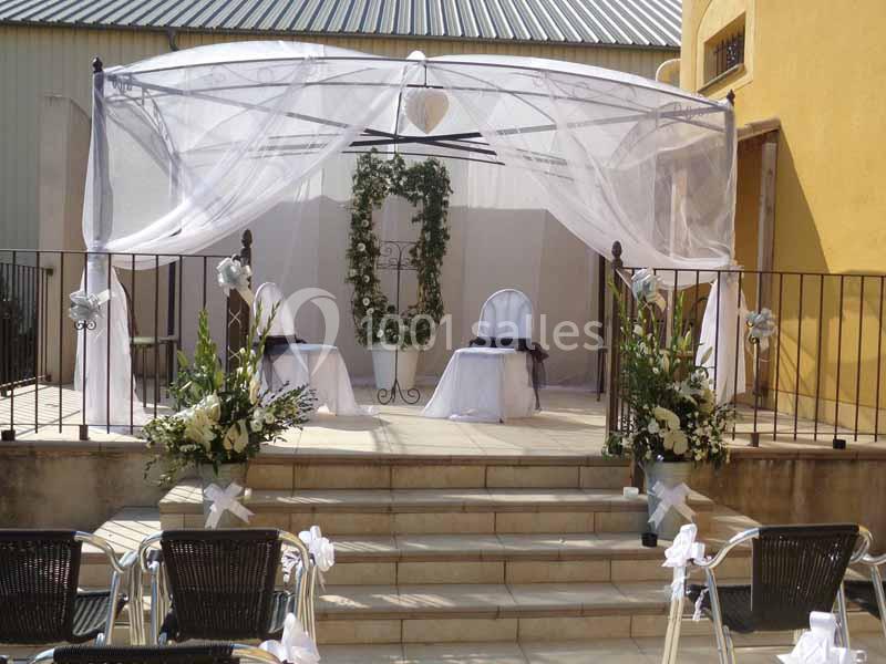 Structure de cérémonie en extérieur avec arche décorée de fleurs, chaises blanches et voilage sous une pergola.