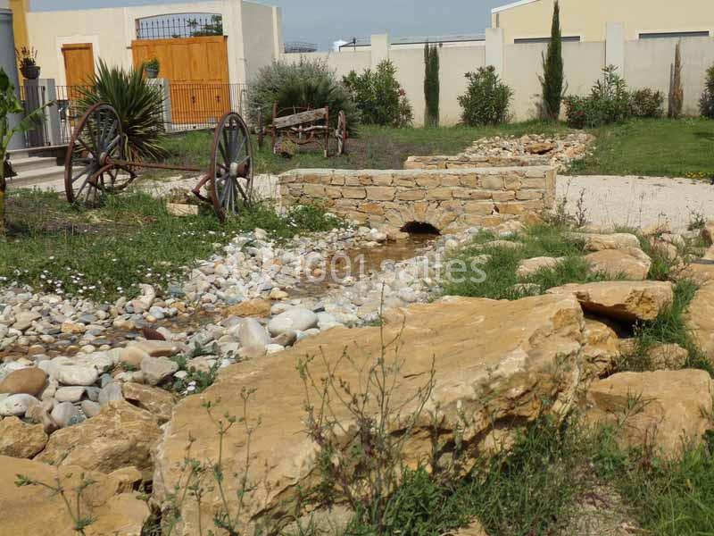 Un jardin avec un ruisseau artificiel, des pierres, des plantes et des éléments décoratifs comme des roues en bois.