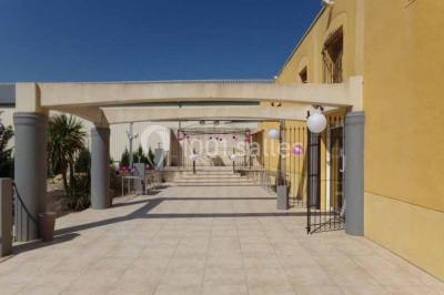 Cour extérieure avec végétation tropicale, terrasse ensoleillée, murs jaunes et mobilier en bois près d'une pergola.