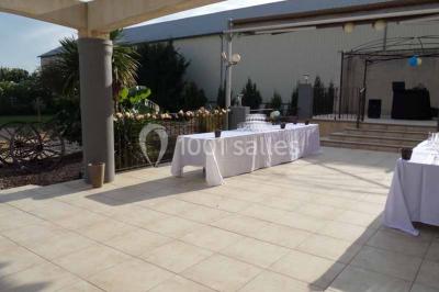Cour extérieure avec végétation tropicale, terrasse ensoleillée, murs jaunes et mobilier en bois près d'une pergola.