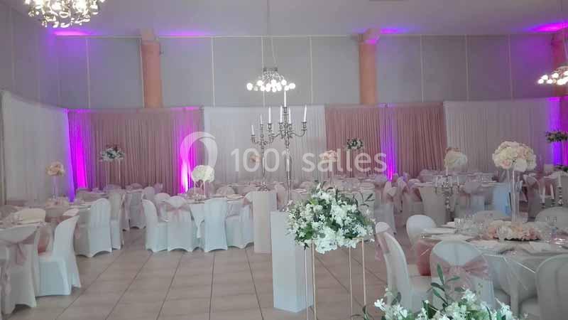 Salle de réception décorée avec des tables rondes, chaises blanches et centres de table fleuris sous un éclairage violet.