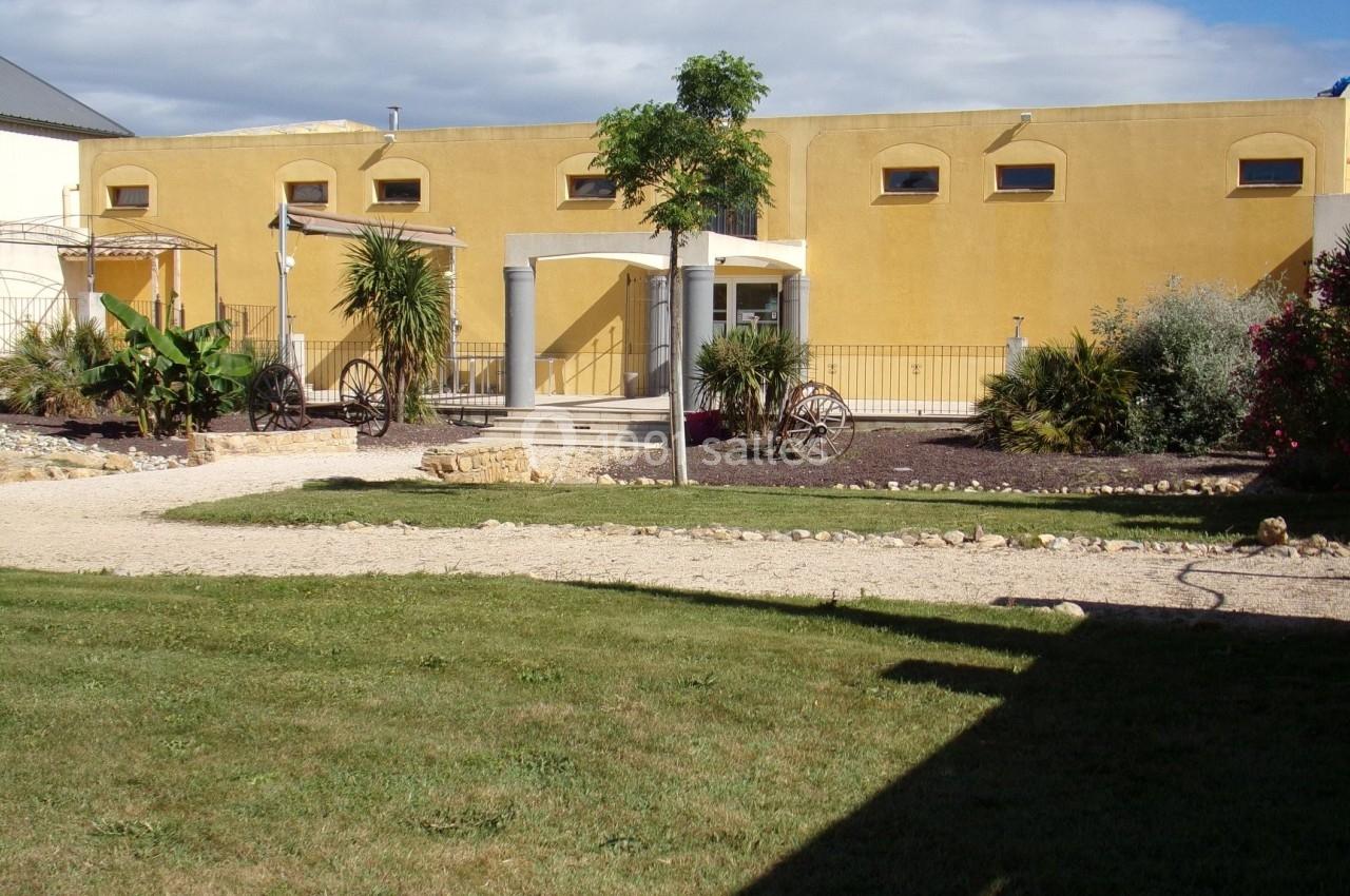 Bâtiment jaune avec des arches, entouré de végétation, d'une allée en gravier et d'une pelouse au premier plan.