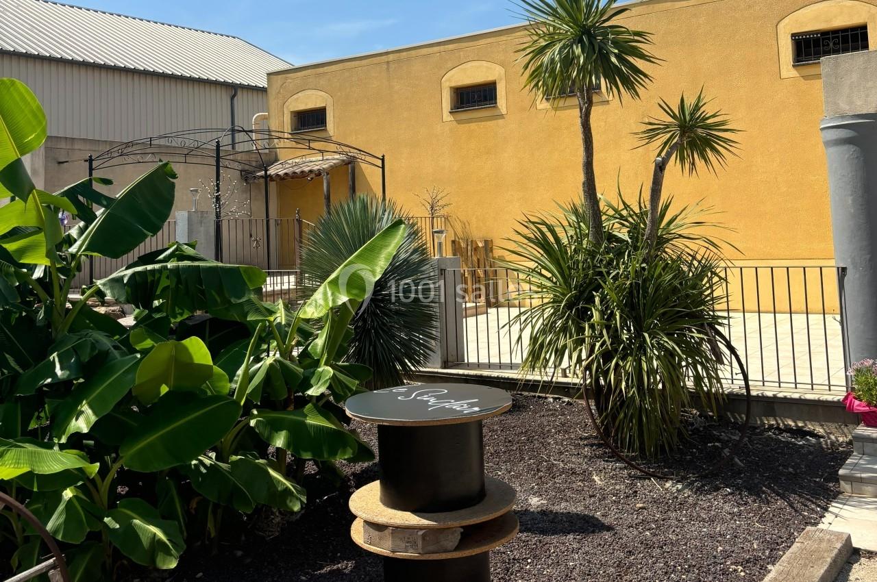 Cour extérieure avec végétation tropicale, terrasse ensoleillée, murs jaunes et mobilier en bois près d'une pergola.
