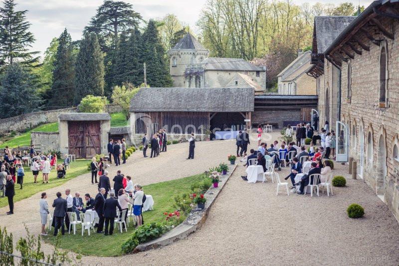 Personnes rassemblées dans la cour d'un domaine rural avec bâtiments en pierre, arbres et pelouse en arrière-plan.