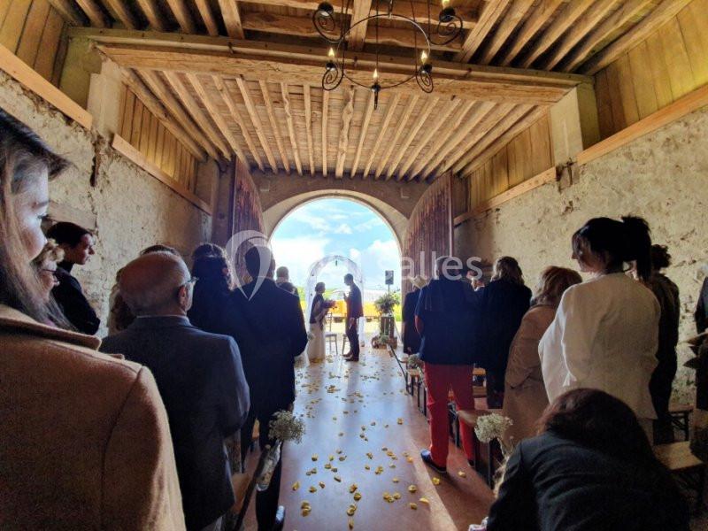 Cérémonie de mariage dans une grange avec des invités assis, vue sur l'extérieur ensoleillé à travers une grande ouverture.