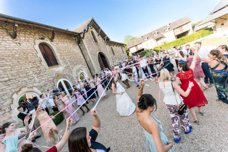 Des invités entourent une mariée en extérieur, tenant des rubans colorés tendus dans une ambiance festive.