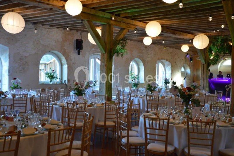 Salle de réception décorée avec des tables rondes, des chaises en bois, des fleurs et des lanternes suspendues.