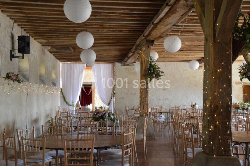 Salle de réception rustique décorée de guirlandes lumineuses, lanternes suspendues et tables dressées avec fleurs.