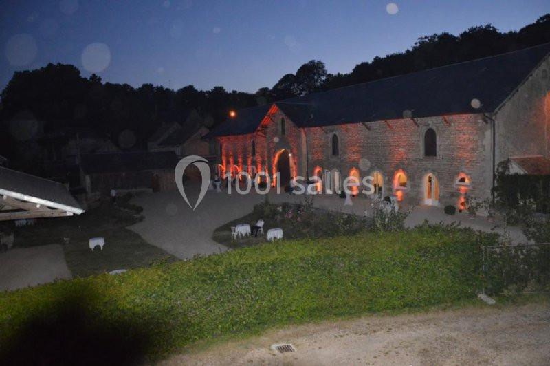 Bâtiment en pierre éclairé de lumières orangées au crépuscule, entouré de végétation et d'une cour.