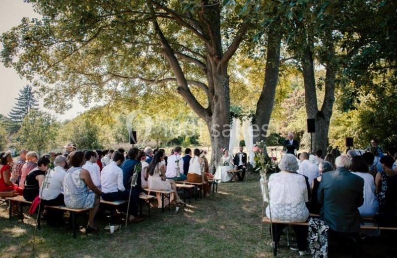 Cérémonie en plein air avec des invités assis sous de grands arbres, face à un officiant et un couple.