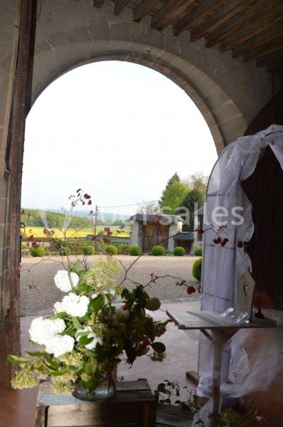 Vue depuis une arche en pierre sur une cour avec des fleurs en premier plan et un paysage champêtre en arrière-plan.
