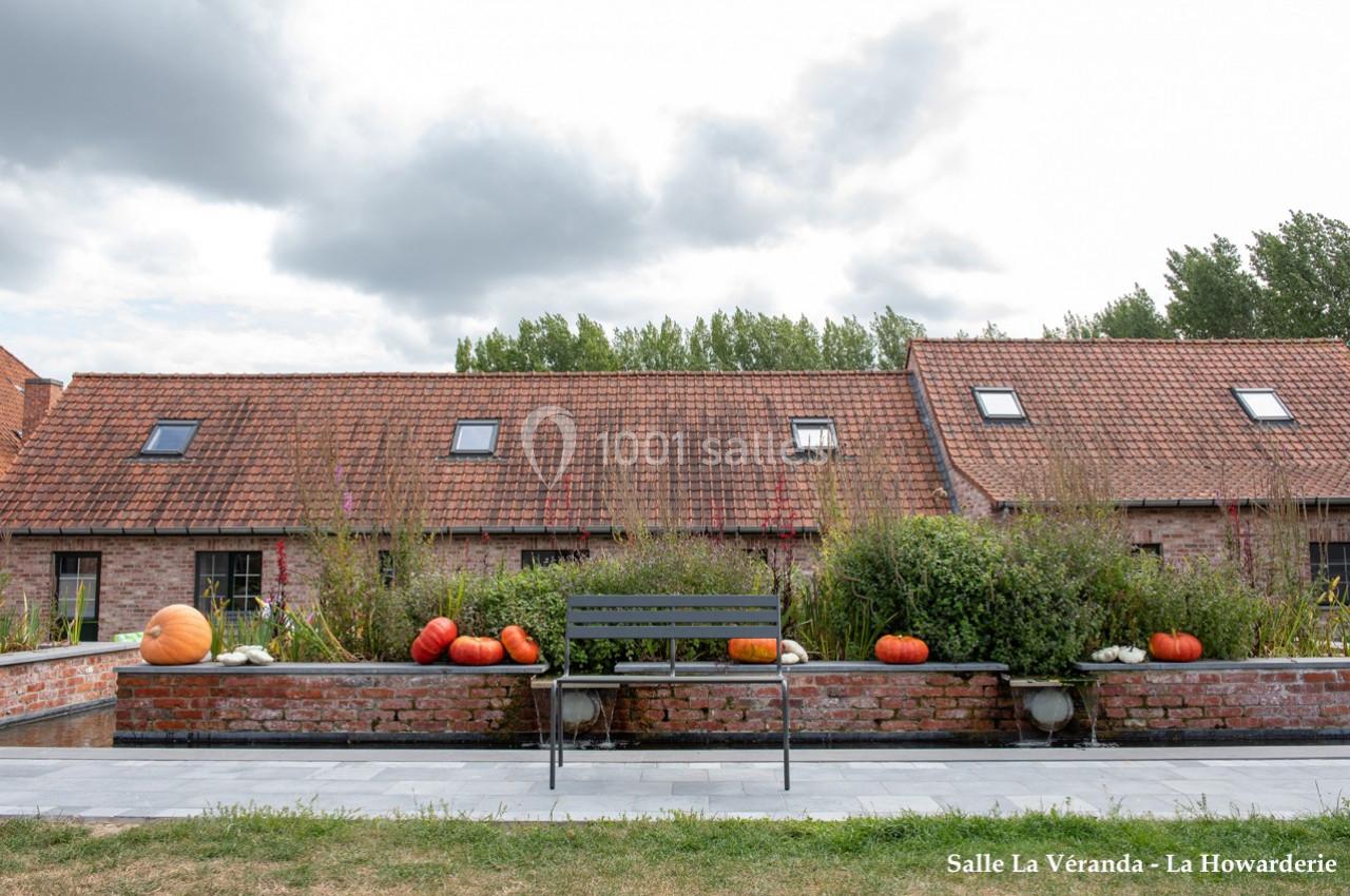 Banc en métal devant un muret décoré de citrouilles, avec des bâtiments en briques et un ciel nuageux en arrière-plan.