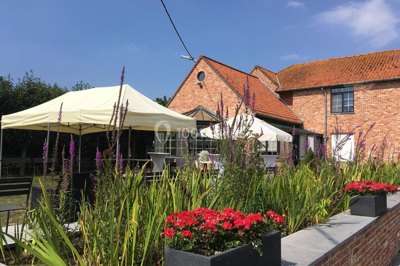 Jardin avec fleurs colorées, terrasse aménagée sous tonnelles blanches, et bâtiment en briques rouges sous ciel bleu.
