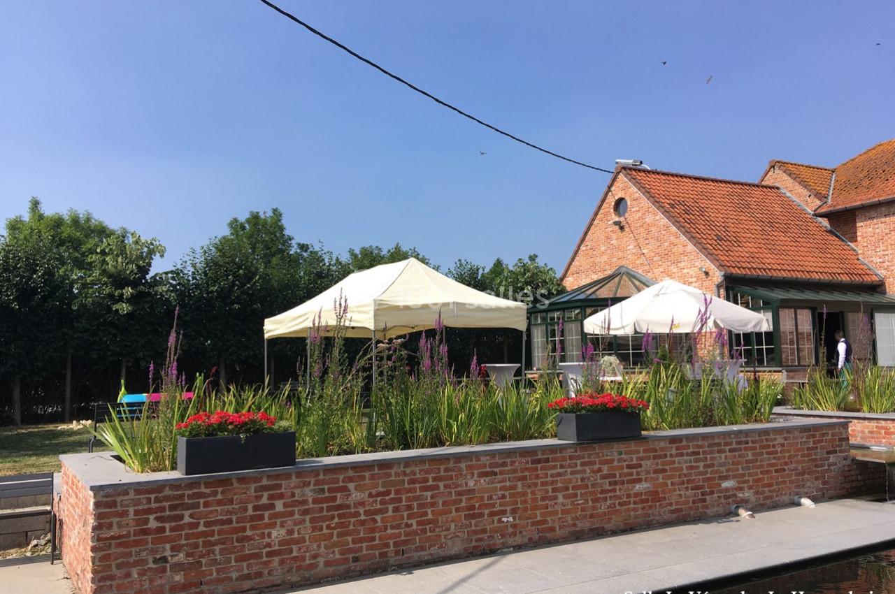 Terrasse ensoleillée avec parterres de fleurs, parasols blancs et bâtiment en briques rouges à l'arrière-plan.