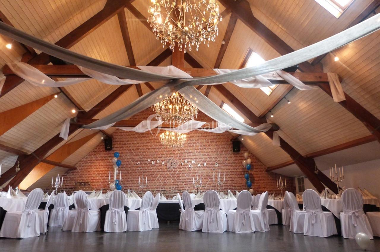 Salle de réception avec plafond en poutres apparentes, tables rondes dressées et chaises blanches, décorée de guirlandes et…