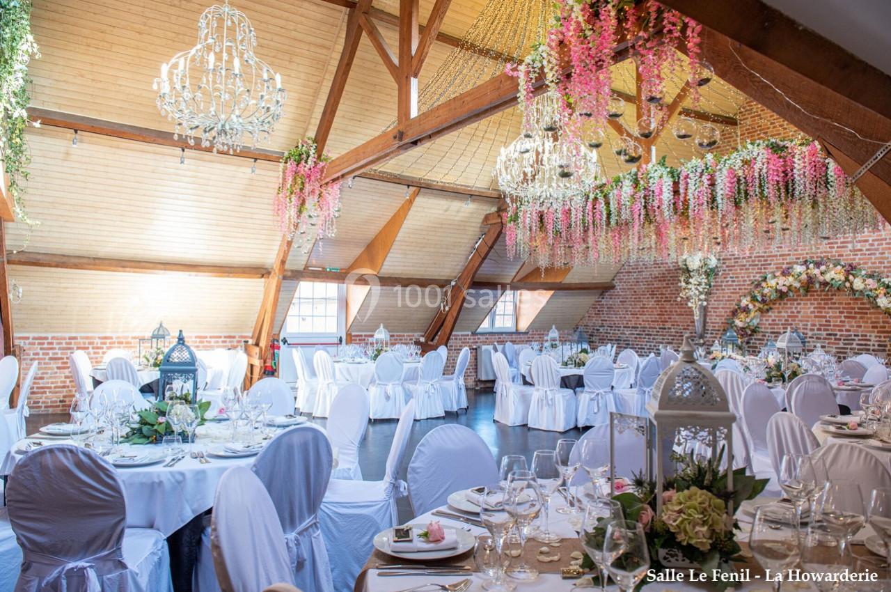 Salle de réception lumineuse avec tables rondes dressées, chaises blanches et décorations florales suspendues.