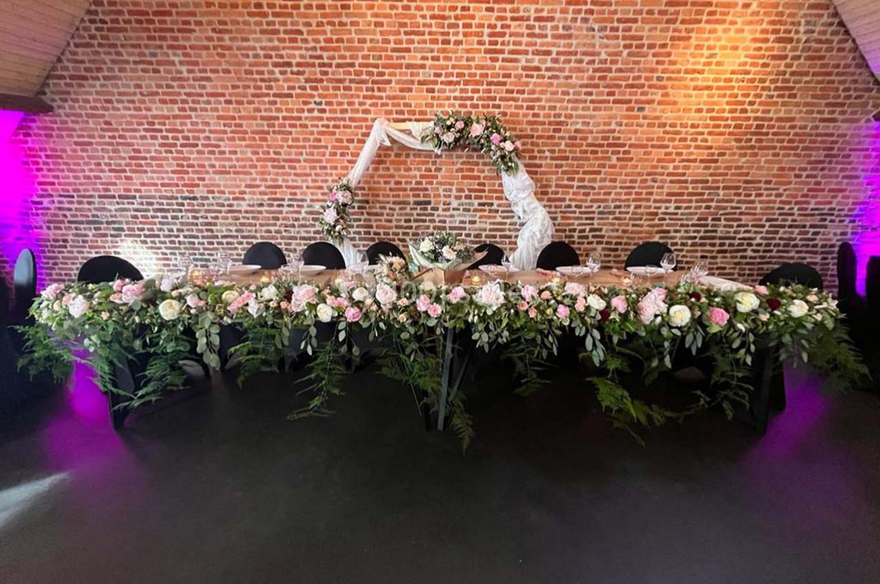 Table décorée pour un événement avec des fleurs roses et blanches, devant un mur de briques éclairé en lumière violette.