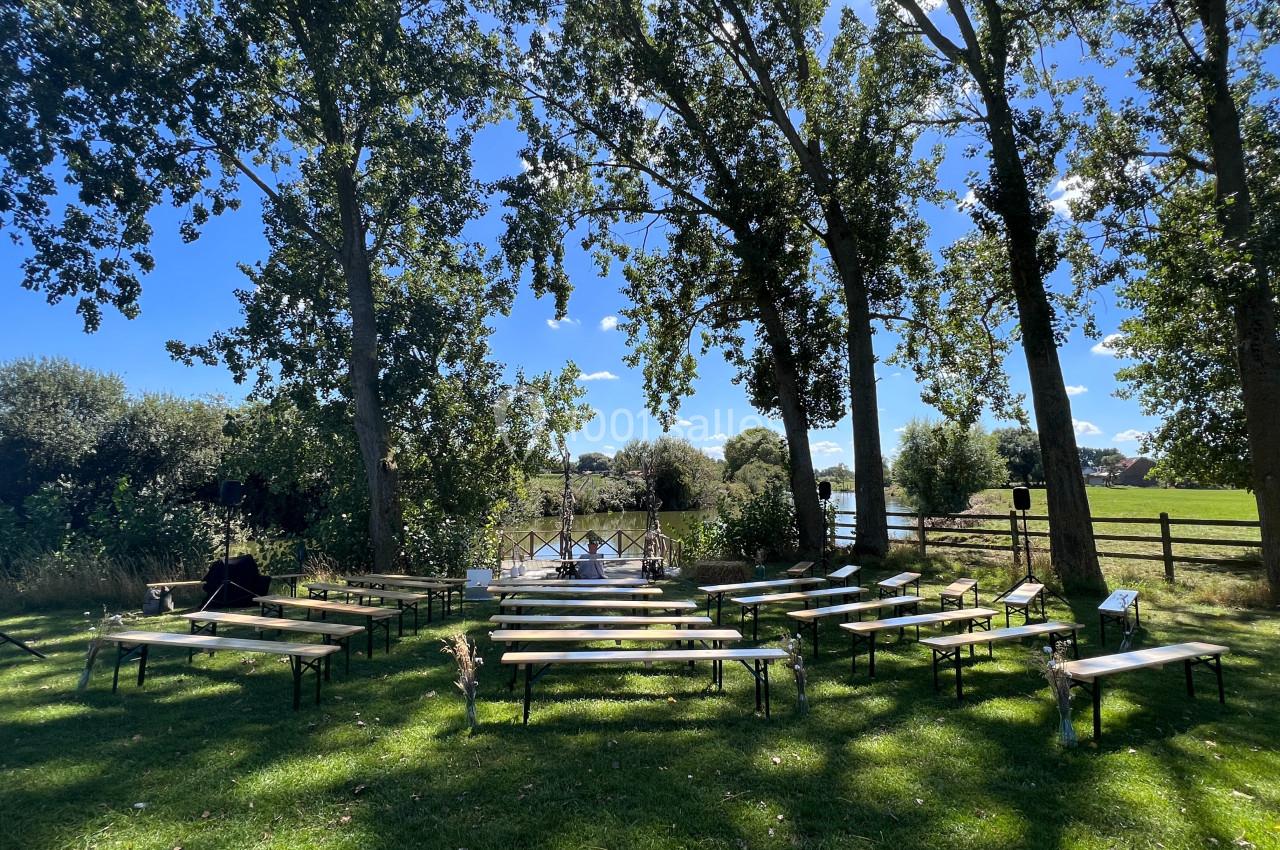 Chaises et bancs disposés en extérieur sous des arbres, face à un point d'eau, par une journée ensoleillée.