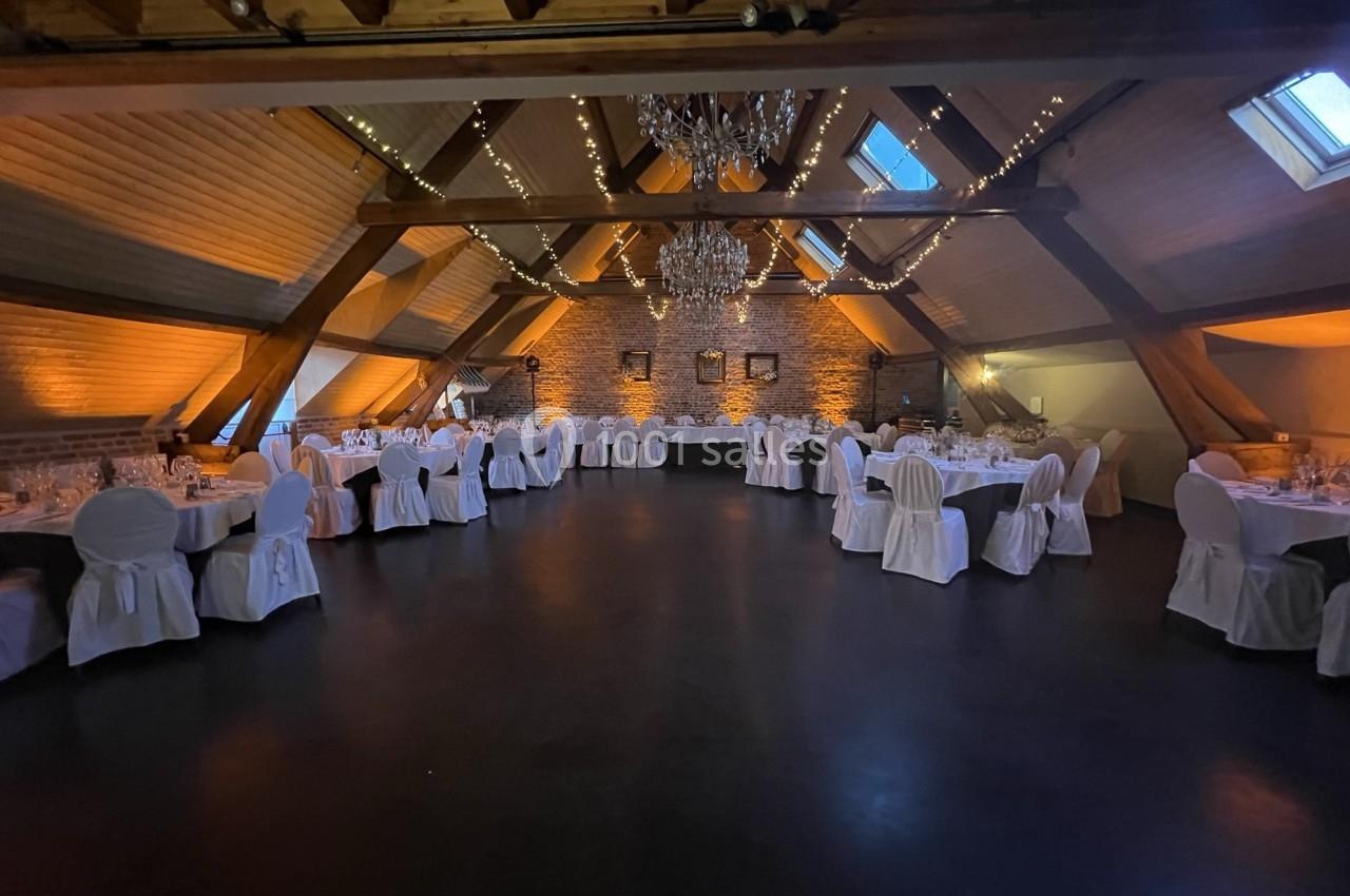 Salle de réception sous combles avec tables rondes dressées, chaises blanches et éclairage chaleureux.