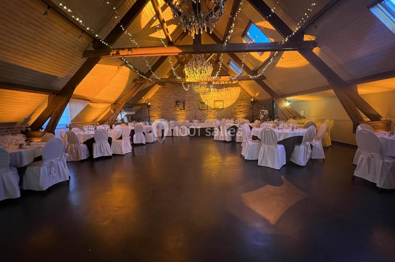 Salle de réception spacieuse avec des tables rondes dressées, éclairage chaleureux et poutres apparentes en bois.