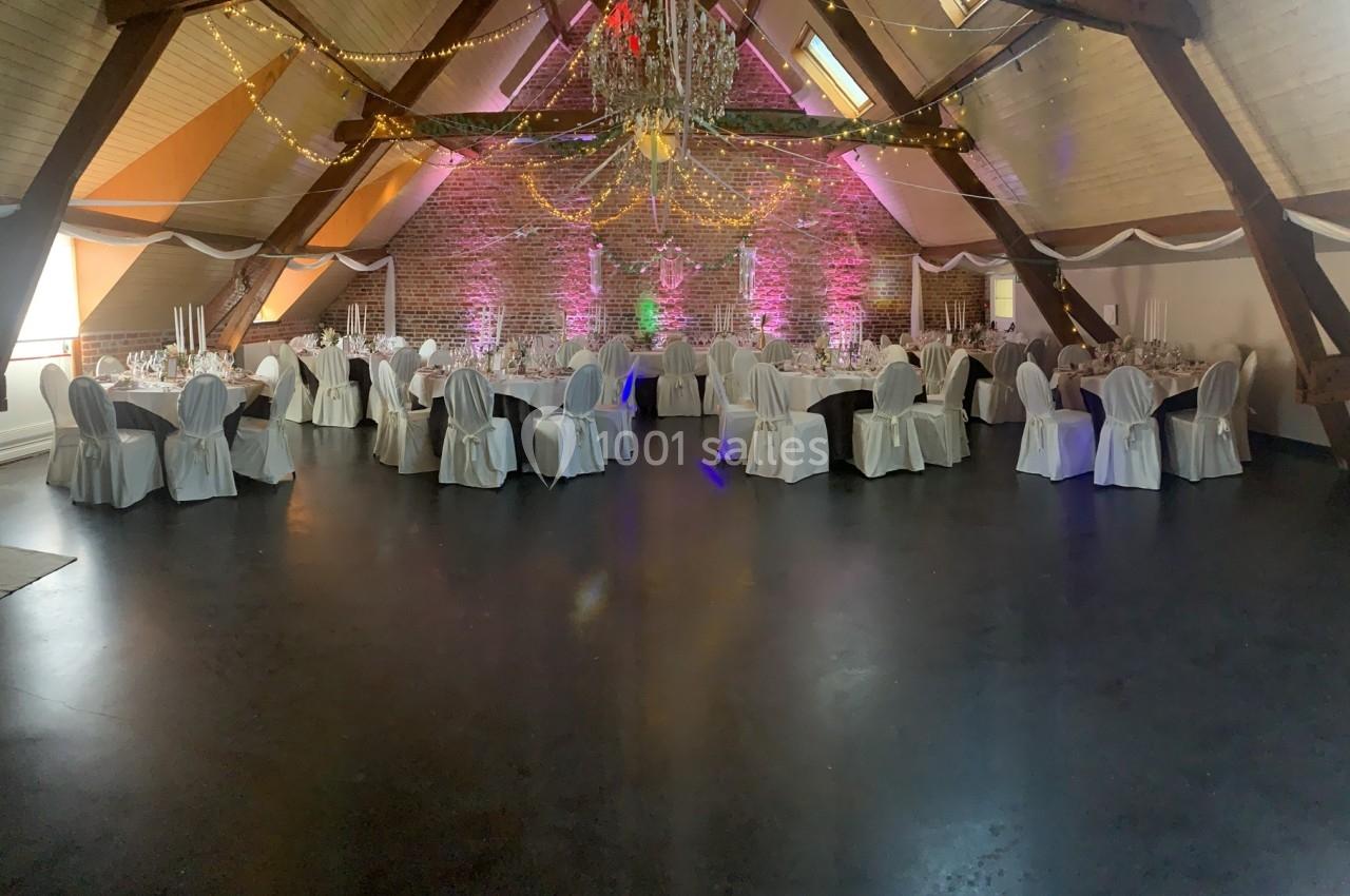 Salle de réception décorée avec des tables rondes, des chaises blanches et un éclairage chaleureux sous une charpente en…