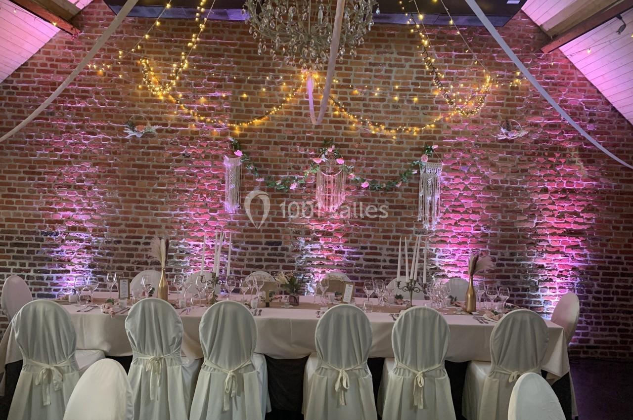 Salle décorée pour un événement avec une table dressée, des chaises blanches, guirlandes lumineuses et un mur de briques.