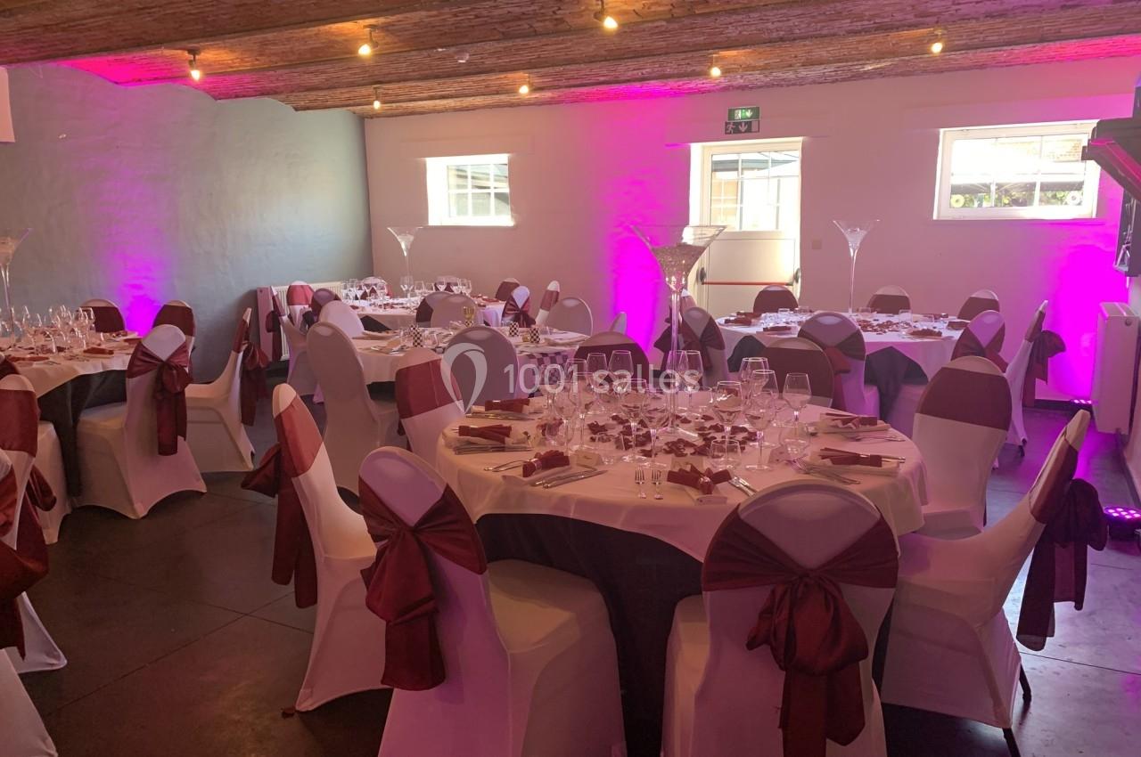 Salle de réception décorée avec des tables rondes dressées, nappes blanches, chaises ornées de nœuds rouges et éclairage…