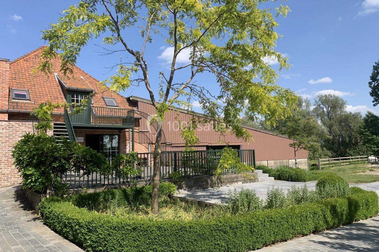 Bâtiment en briques et métal avec terrasse, entouré de verdure et d'un chemin pavé sous un ciel bleu.
