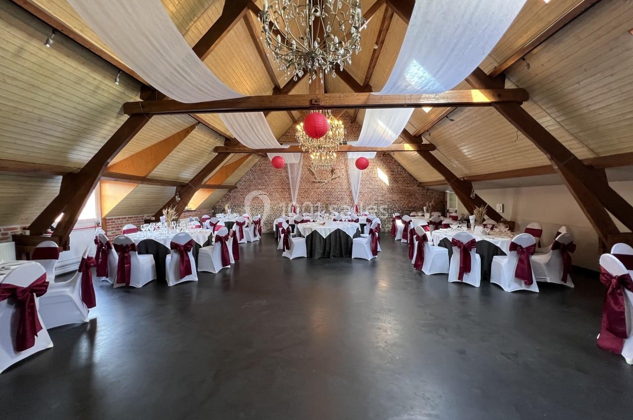 Salle de réception décorée avec des tables rondes, nappes blanches, chaises ornées de nœuds bordeaux et lustres suspendus.
