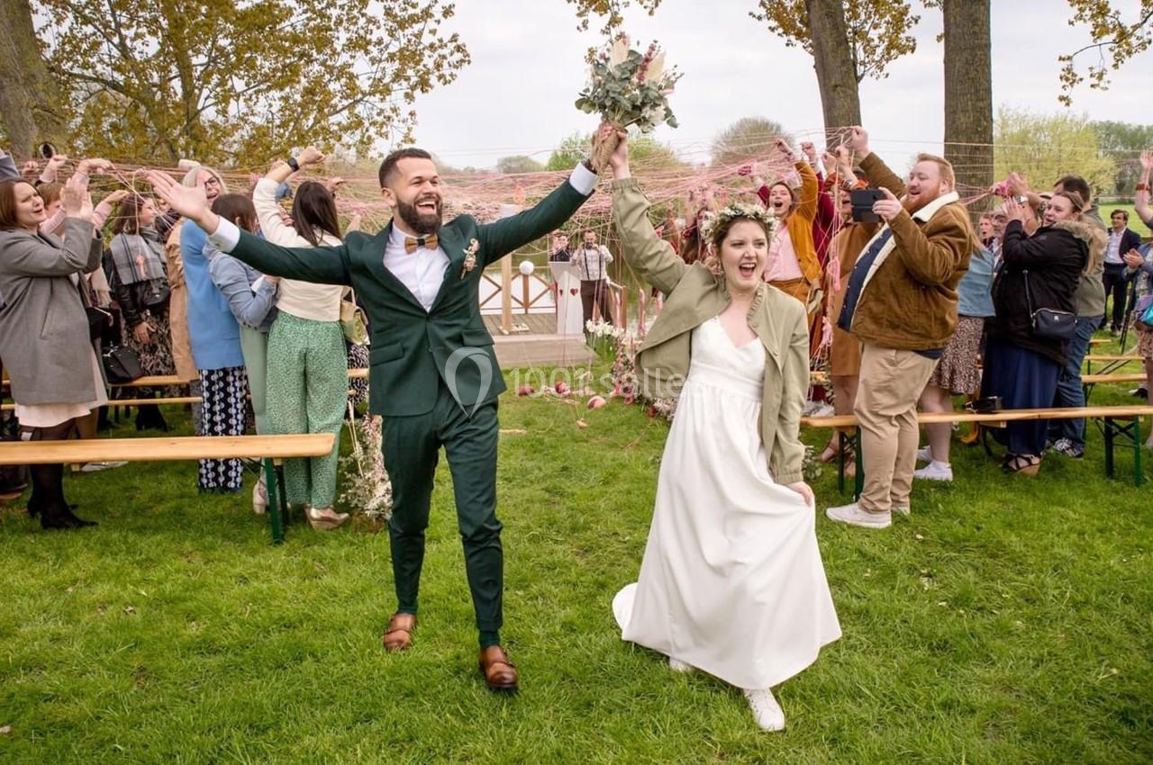 Un couple souriant en tenue de mariage marche sur une pelouse, entouré d'invités célébrant avec enthousiasme.